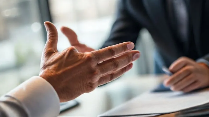 Photorealistic, professional photography, 8K, cinematic lighting, sharp focus, depth of field, shot on a high-end DSLR. A close-up of a sales professional's hand gesturing gently during a conversation, conveying empathy and openness. The prospect's hand is visible in the foreground, perhaps holding a pen, indicating active listening and consideration. A subtle blur of a modern office environment in the background.